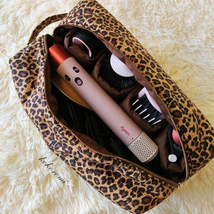 May include: A leopard print zippered travel case containing a pink Dyson hair styling tool with a metallic orange top, a round brush attachment, and other hair styling accessories. The word kokoromi is printed in black on the lower left side.