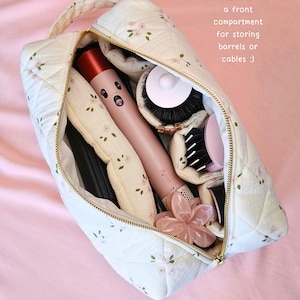 May include: A quilted cream-coloured storage bag with a delicate pink floral pattern, featuring a gold zip and a top handle. The bag contains a pink hair styling tool, various round brush attachments, and a flower-shaped hair clip. Text reads: Every design features a front compartment for storing barrels or cables :)