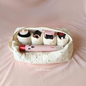 May include: A quilted cream-coloured fabric storage pouch with a delicate floral pattern, featuring a gold zip and internal compartments holding a pink Dyson hair styling tool and four various black and pink hair styling attachments.