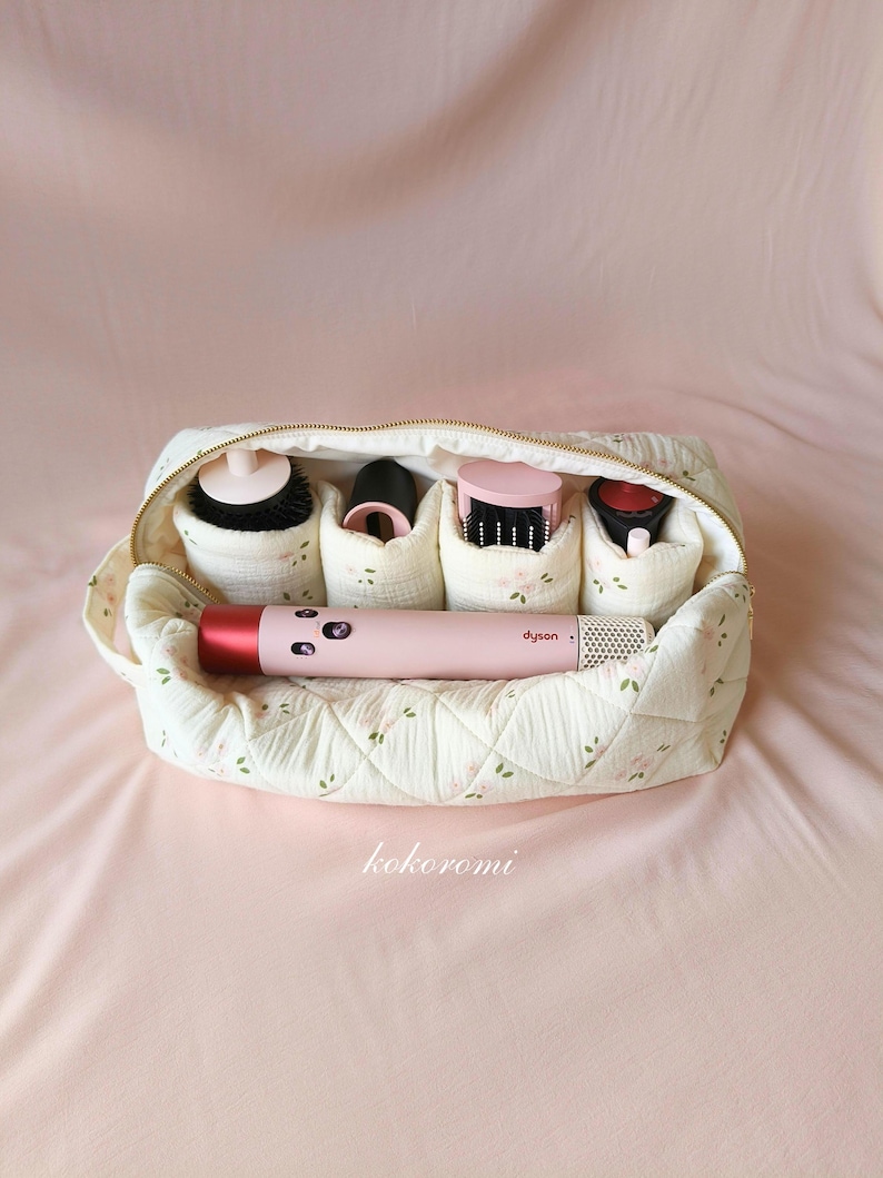 May include: A quilted cream-coloured fabric storage pouch with a delicate floral pattern, featuring a gold zip and internal compartments holding a pink Dyson hair styling tool and four various black and pink hair styling attachments.
