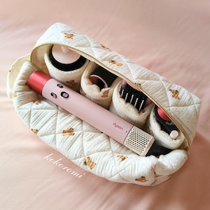 May include: A quilted cream-coloured storage pouch with a brown teddy bear pattern and gold zip, containing a light pink Dyson hair styling tool and several black and pink hair styling attachments organised in individual interior compartments.