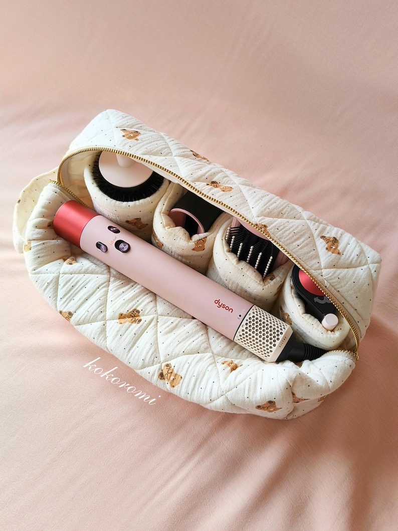 May include: A quilted cream-coloured storage pouch with a brown teddy bear pattern and gold zip, containing a light pink Dyson hair styling tool and several black and pink hair styling attachments organised in individual interior compartments.