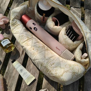 May include: A quilted cream-coloured cosmetic bag with a gold floral pattern containing a pink Dyson hair styling tool, various hair brushes, and attachments. Outside the bag are a bottle of Gisou hair oil, a tube of Beyond hand cream, and a scrunchie.