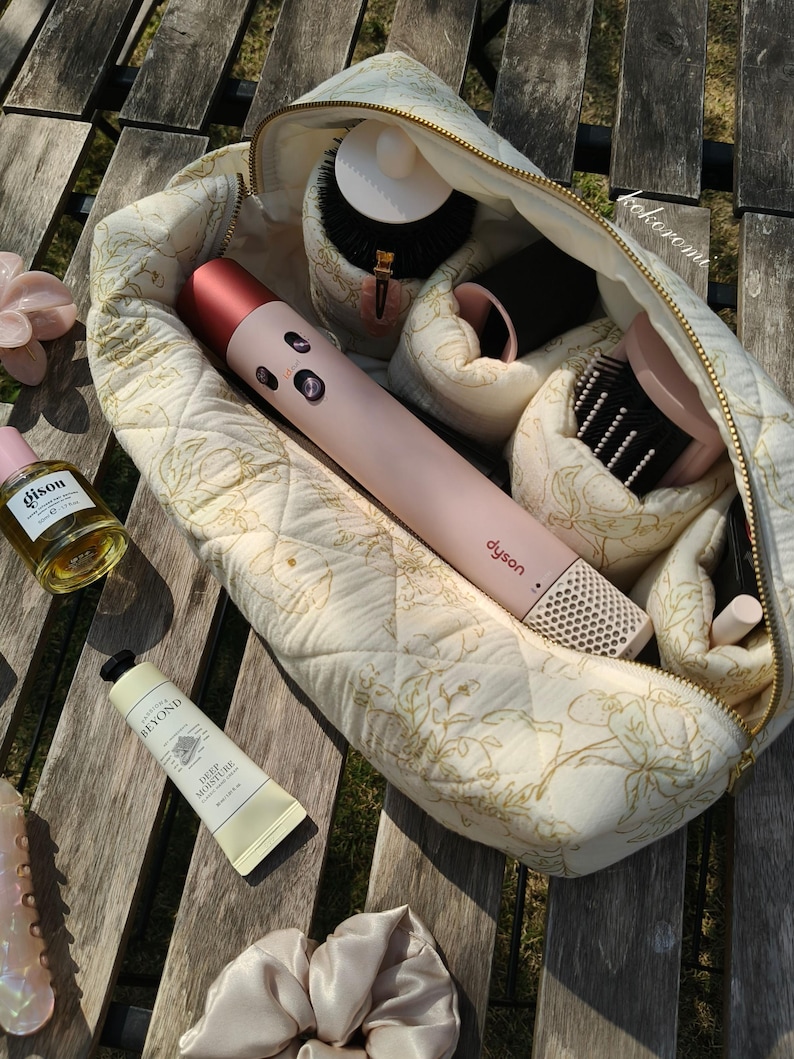 May include: A quilted cream-coloured cosmetic bag with a gold floral pattern containing a pink Dyson hair styling tool, various hair brushes, and attachments. Outside the bag are a bottle of Gisou hair oil, a tube of Beyond hand cream, and a scrunchie.