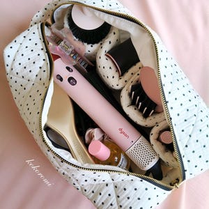 May include: A white quilted travel bag with black polka dots and a gold zip, filled with hair styling tools including a pink Dyson hair styler, a beige hairbrush, a small bottle with a pink cap, and various hair accessories tucked inside.
