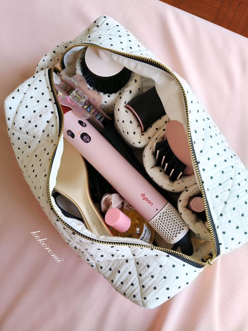 May include: A white quilted travel bag with black polka dots and a gold zip, filled with hair styling tools including a pink Dyson hair styler, a beige hairbrush, a small bottle with a pink cap, and various hair accessories tucked inside.