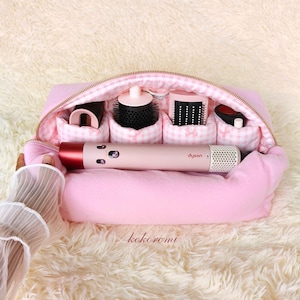 Puede incluir: Un estuche de almacenamiento rosa suave con cremallera y un forro interior de cuadros vichy en rosa claro y blanco. El estuche contiene una herramienta de peinado Dyson de color rosa y cuatro compartimentos internos con accesorios negros.