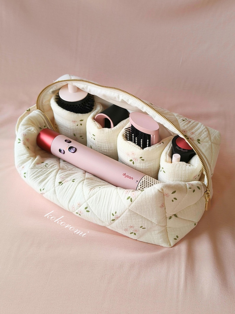 May include: A quilted cream-coloured storage pouch with a small pink floral pattern and the text "kokoromi" printed on the front. The open pouch contains a pink Dyson hair styling tool and several black and pink hair styling attachments.