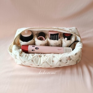 May include: A cream-coloured quilted travel case with a small floral pattern, featuring a gold zip and internal compartments holding a pink Dyson hair styling tool and various black and pink attachments, with the word kokoromi printed below.