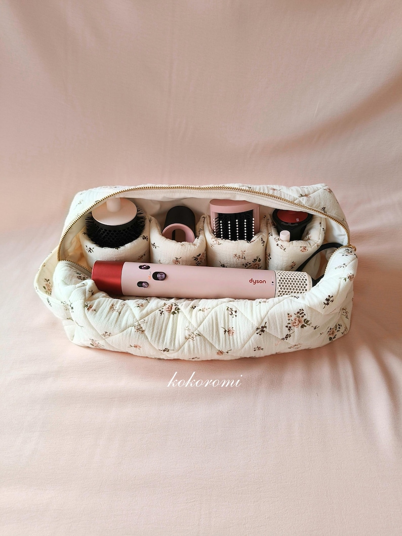 May include: A cream-coloured quilted travel case with a small floral pattern, featuring a gold zip and internal compartments holding a pink Dyson hair styling tool and various black and pink attachments, with the word kokoromi printed below.