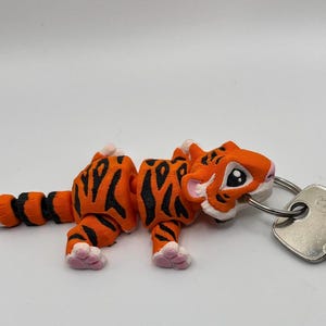 Op de afbeelding: Een 3D-geprinte oranje en zwarte tijger sleutelhanger met een wit gezicht en roze pootjes. De tijger is bevestigd aan een zilveren sleutelring.