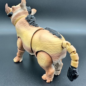 Könnte beinhalten: Ein bewegliches Stier-Figürchen in Braun- und Beigetönen mit schwarzen Akzenten auf Rücken und Schwanz. Das Spielzeug steht auf allen Vieren und zeigt sein detailliertes Design und die segmentierten Gelenke. Die Figur hat eine strukturierte Oberfläche.
