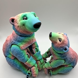 Könnte beinhalten: Zwei bewegliche Bärenfiguren in Regenbogenfarben. Die Bären haben schwarze Nasen und sitzen. Der größere Bär befindet sich neben einem kleineren Bären, beide mit freundlichem Gesichtsausdruck.