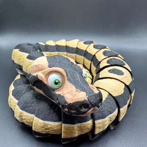 Peut inclure: Un jouet serpent imprim&eacute; en 3D avec des rayures noires et beiges et de grands yeux verts. Le serpent est enroul&eacute; en cercle, la t&ecirc;te face &agrave; l'appareil photo.