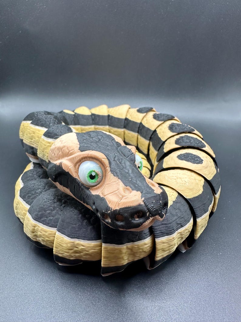 Peut inclure: Un jouet serpent imprim&eacute; en 3D avec des rayures noires et beiges et de grands yeux verts. Le serpent est enroul&eacute; en cercle, la t&ecirc;te face &agrave; l'appareil photo.
