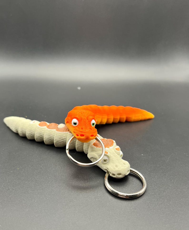 May include: Two orange and white worm-shaped keyrings with a silver key ring. The orange worm has a face with eyes and a mouth. The white worm has a face with eyes and a mouth.