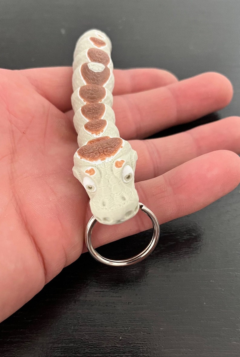May include: A white and brown plastic snake keyring with a silver ring. The snake has a textured body and a detailed head with eyes.