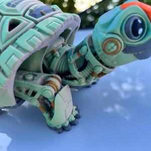 Peut inclure: Une tortue robotique verte et grise avec des accents orange. La tortue a un corps mécanique détaillé et une grosse tête ronde avec un seul œil.