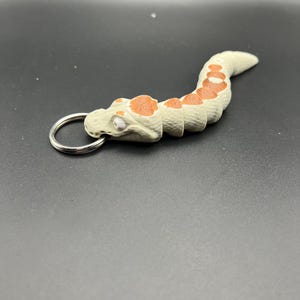 May include: A beige and orange snake-shaped keyring with a silver ring. The snake has a textured surface and a small white eye.