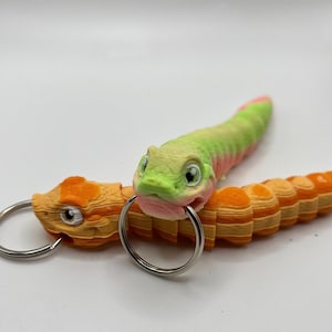 Könnte beinhalten: Zwei bewegliche Schlangen-Schlüsselanhänger. Einer ist orange und gelb, der andere grün, rosa und gelb. Jeder hat einen silbernen Schlüsselring an seinem Maul befestigt. Die Schlangen haben detaillierte Augen und segmentierte Körper.