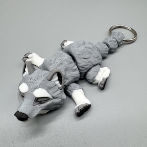 Könnte beinhalten: Ein 3D-gedruckter, beweglicher Wolf-Schlüsselanhänger mit grauer und weißer Schichtstruktur. Der Wolf ist in liegender Position mit geschlossenen Augen, weißen Pfoten und einem segmentierten Schwanz an einem runden silbernen Metallring dargestellt.