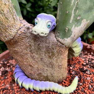 Può includere: Un giocattolo a forma di serpente segmentato di colore viola e verde con grandi occhi blu, che spunta da dietro una pianta di cactus.