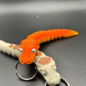 May include: Two orange and white snake-like keyrings with googly eyes. The orange keyring is curled into a C shape and the white keyring is straight. Both keyrings have a silver ring attached.