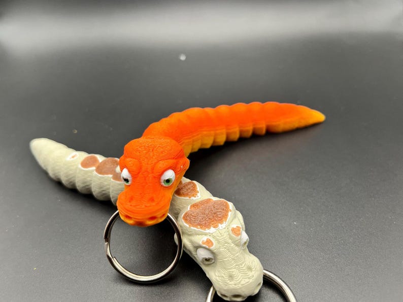 May include: Two orange and white snake-like keyrings with googly eyes. The orange keyring is curled into a C shape and the white keyring is straight. Both keyrings have a silver ring attached.