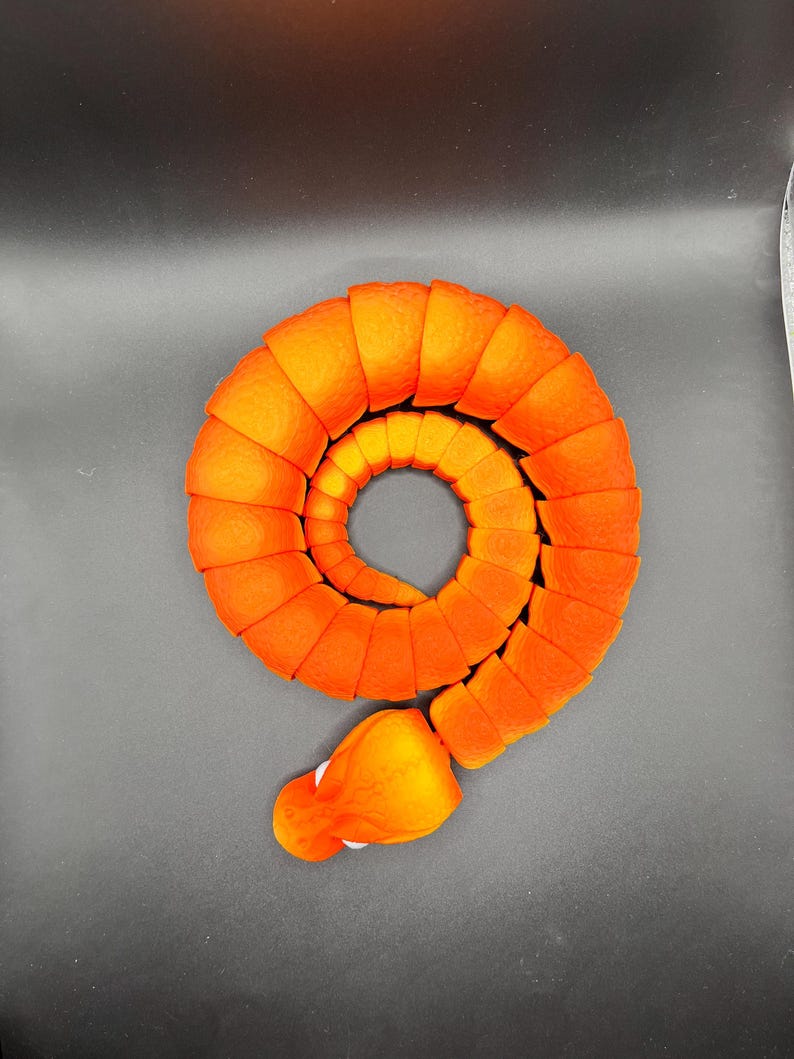 Peut inclure: Un serpent de jouet orange et jaune segment&eacute; enroul&eacute; en cercle. Le serpent a une t&ecirc;te arrondie avec des yeux blancs.