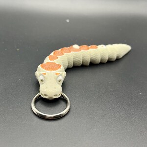 May include: A white and brown snake keyring with a silver ring. The snake has a segmented body and a detailed head with eyes.
