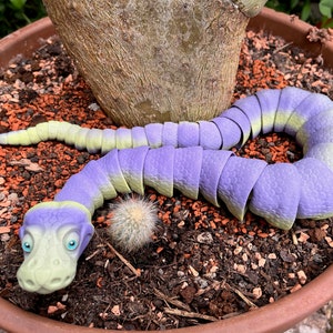 Peut inclure: Un jouet en forme de serpent segment&eacute;, de couleur violette et verte, est enroul&eacute; autour d'une plante en pot. Le serpent a une grosse t&ecirc;te avec une expression amicale et de grands yeux bleus.