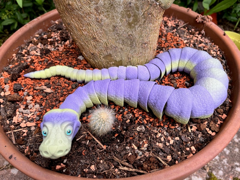 Peut inclure: Un jouet en forme de serpent segment&eacute;, de couleur violette et verte, est enroul&eacute; autour d'une plante en pot. Le serpent a une grosse t&ecirc;te avec une expression amicale et de grands yeux bleus.