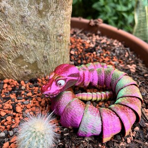 Peut inclure: Un jouet serpent segment&eacute; rose et vert avec un gros &oelig;il, enroul&eacute; en cercle sur un lit de gravier brun et rouge. Une petite plante de cactus se trouve au premier plan.