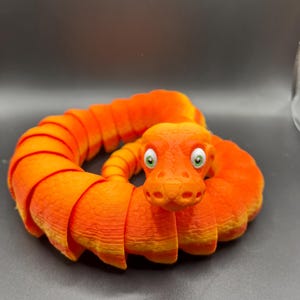 Peut inclure: Un jouet en forme de serpent segment&eacute; orange et jaune avec des yeux verts. Le serpent est enroul&eacute; en cercle.