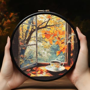 May include: A cross-stitch embroidery in a black hoop depicts an autumn scene. The image shows a window overlooking a tree with yellow leaves, a cup of coffee, and fallen leaves. The embroidery is held by hands.