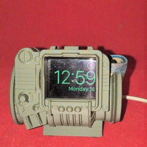 May include: A green 3D printed stand for an Apple Watch. The stand is shaped like a Pip-Boy from the Fallout video game series. The watch face shows the time 12:59 and the date Monday 14.