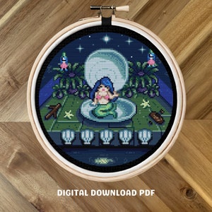 May include: A cross-stitch pattern featuring a mermaid sitting in a seashell under a full moon. The pattern is surrounded by a border of seashells and stars. The pattern is in a wooden embroidery hoop.