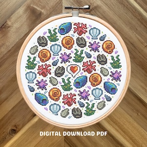 May include: A round cross-stitch pattern featuring a variety of sea-themed icons, including seashells, starfish, coral, seaweed, and rocks. The pattern is in a white hoop with a wooden frame.