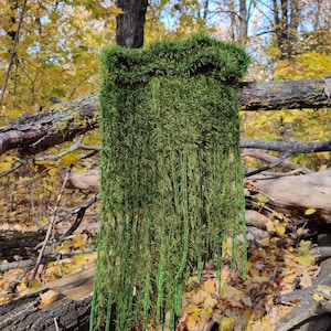 Könnte beinhalten: Ein langer, grüner, fransiger Schal, der an einem Baumzweig in einem Wald hängt. Der Schal ist aus einem weichen, flauschigen Material gefertigt und hat einen langen Fransen, der bis zum Boden reicht. Der Schal ist in einem leuchtenden Grün gehalten und eine schöne Ergänzung zu jedem Outfit.