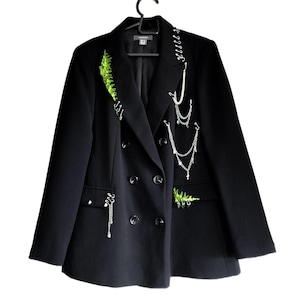 Pode incluir: Um blazer preto de abotoamento duplo com correntes decorativas prateadas e detalhes bordados em verde. O blazer apresenta lapelas entalhadas, quatro botões e está pendurado em um cabide preto.