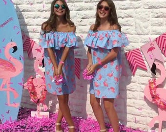May include: Two people wearing matching blue off-the-shoulder dresses with pink flamingo print. The dresses have a ruffled neckline and short sleeves. They are standing in front of a white brick wall with pink confetti on the ground.