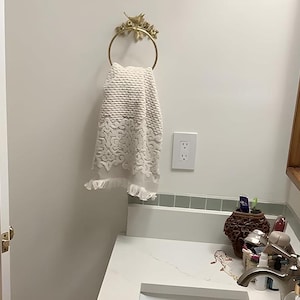 Puede incluir: Un toallero dorado con un dise&ntilde;o decorativo de p&aacute;jaros y flores sostiene una toalla blanca con un patr&oacute;n texturizado y ribete de encaje. La toalla cuelga en un ba&ntilde;o con una encimera y una pared blancas.