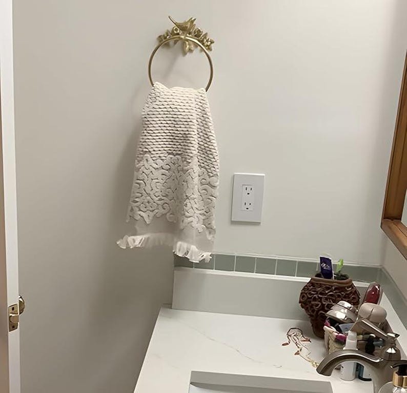 Puede incluir: Un toallero dorado con un dise&ntilde;o decorativo de p&aacute;jaros y flores sostiene una toalla blanca con un patr&oacute;n texturizado y ribete de encaje. La toalla cuelga en un ba&ntilde;o con una encimera y una pared blancas.