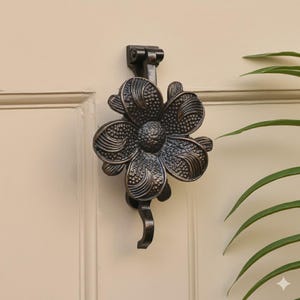 K&ouml;nnte beinhalten: Bronzefarbener T&uuml;rklopfer in Blumenform. Der T&uuml;rklopfer ist an einer cremefarbenen T&uuml;r befestigt. Die Blume hat detaillierte Bl&uuml;tenblattmuster und einen gebogenen Griff. Ein dekoratives Element f&uuml;r jeden Hauseingang.