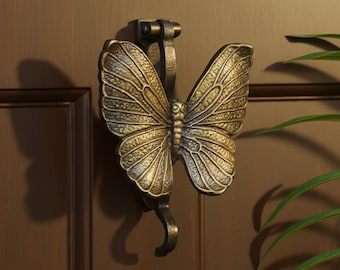 Könnte beinhalten: Ein bronzefarbener Türklopfer in Schmetterlingsform. Die Schmetterlingsflügel sind mit Adernmustern versehen. Der Klopfer ist an einer braunen Tür befestigt. Das Design verleiht jedem Eingang eine dekorative Note.
