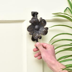 K&ouml;nnte beinhalten: Ein bronzefarbener T&uuml;rklopfer in Form einer Blume. Die Blume hat detaillierte Bl&uuml;tenblattmuster und ist an einer cremefarbenen T&uuml;r angebracht. Eine Hand zieht am Griff des T&uuml;rklopfers. Dekorative Wohnkultur.