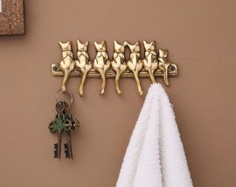 Könnte beinhalten: Ein goldfarbener Metall-Wandhaken mit sechs Katzensilhouetten, die nebeneinander sitzen. Der Haken hält einen Satz Schlüssel und ein weißes Handtuch. Die Katzen sind mit sichtbaren Ohren und Schwänzen detailliert. Der Wandhaken ist an einer braunen Wand befestigt.