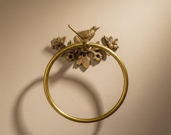 May include: A gold-colored towel ring with a decorative bird and floral design. The ring is circular, and the bird is perched atop a floral arrangement. The fixture is mounted on a beige wall.