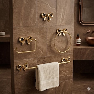 May include: A set of gold-colored bathroom fixtures, including a towel bar, towel ring, toilet paper holder, and a hook. Each piece features a decorative bow design. A white waffle-weave towel hangs on the bar, set against a brown tiled wall.