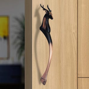 May include: A decorative door handle shaped like a deer. The handle is black and copper, featuring a deer head and a curved body. It's mounted on a light wood door. The handle's design adds a touch of nature.